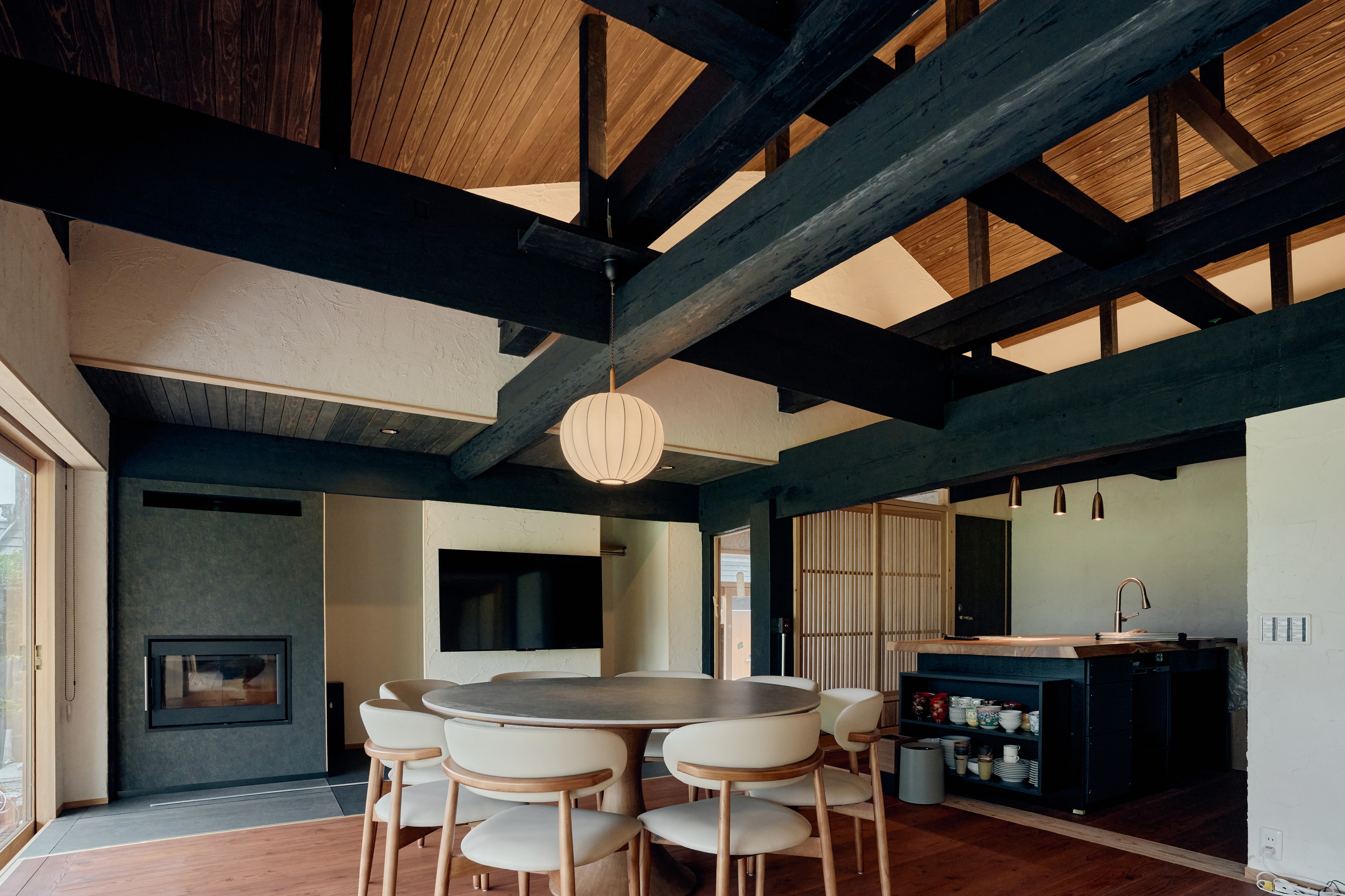 A round dining table beneath exposed timber beams, beside the fireplace and handcrafted kitchen